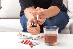 Person lighting a cigarette in front of a table with pills and alcohol on it
