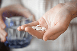 A hand holding pills, with another hand in the background holding a glass of water