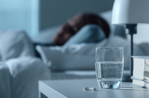 Person sleeping next to a nightstand with a glass of water and pills on it