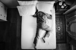 Man in black and white trying to sleep on his bed with a pillow over his head